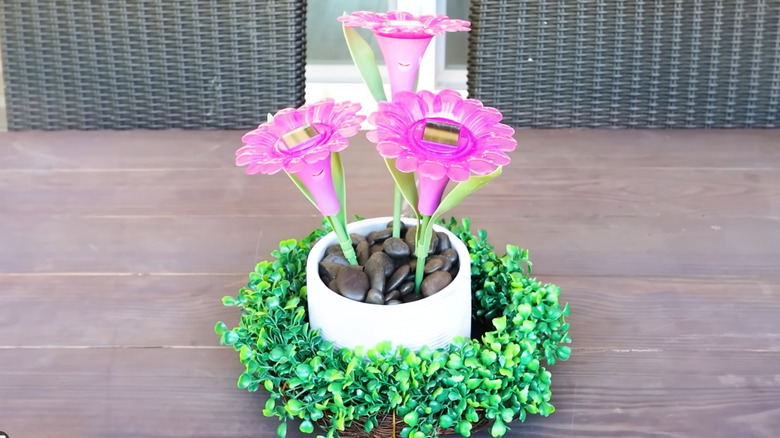 Three purple flowers in a pot with rocks on top and artificial greenery around the base