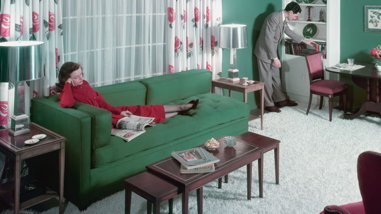 woman reading on a couch in a 1940s living room as a man looks at books on a bookshelf.