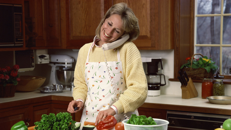 A blond woman from the 1990s, wearing a light yellow sweater and white floral apron, is chopping vegetables and talking on a cream-colored corded phone.