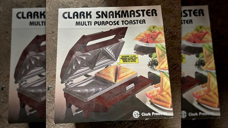 A cardboard box reads "Clark Snakmaster Multi Purpose Toaster" and shows a photo of a sandwich press along with plates of sandwiches, tomatoes, and cut celery.