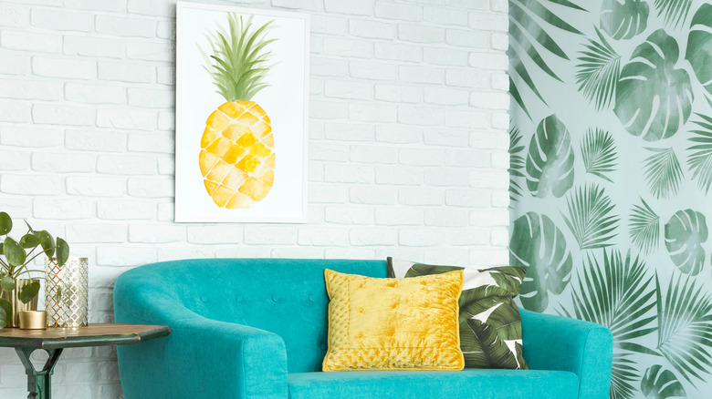 A room with teal couch and framed pineapple art picture above it next to a leaf patterned wallpaper.