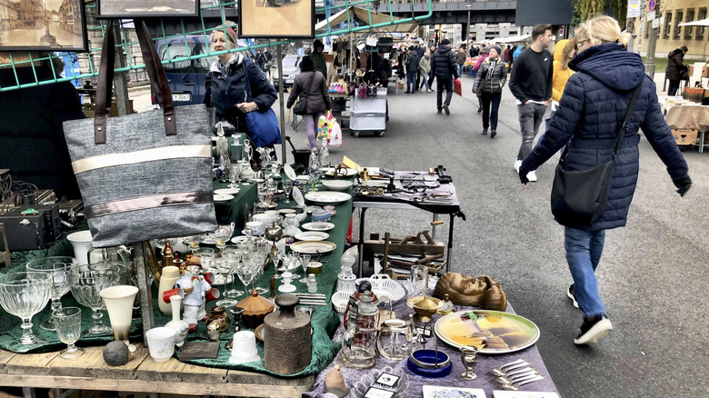 A woman browsing a table covered with items at a flea market while another woman in a puffy blue coat passes by