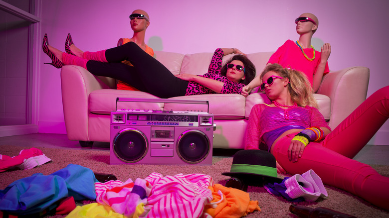 Two women dressed in '80s clothes lounging on a couch surrounded by clothes, mannequins, and a radio
