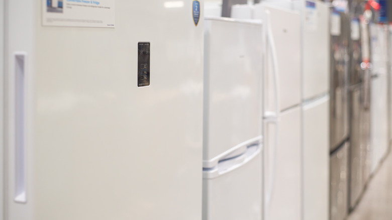 Several white and stainless-steel refrigerators stand in a line in a department store.