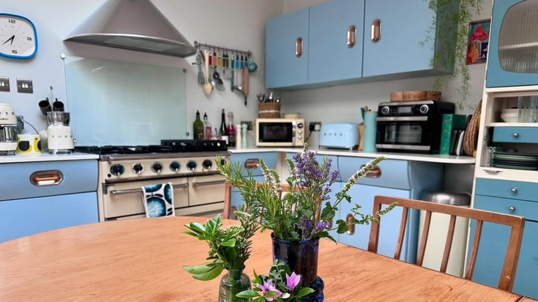 A wooden table with flowers in a vase in front of a 1950s-style kitchen with light blue counters and a gas stove.