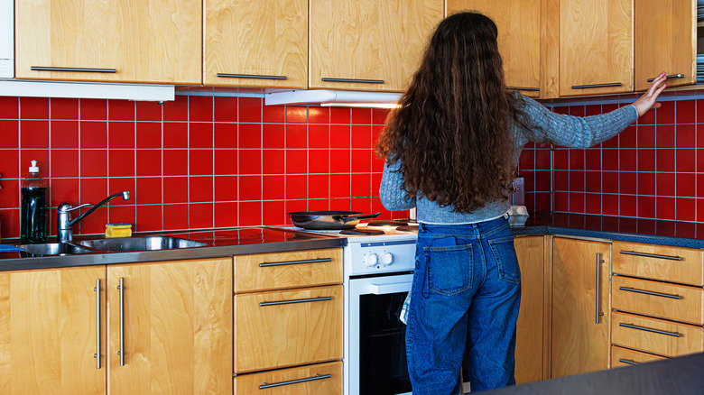 Woman opening cabinet in retro kitchen with slab cabinets and red square tile backsplash
