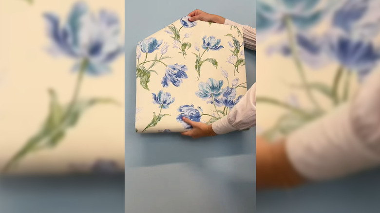 Hands unrolling a sheet of Laura Ashley wallpaper with blue flowers on a white background