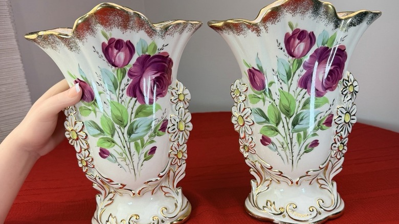 A hand holds one vase in a set of two porcelain fan vases adorned with painted flowers and gold.
