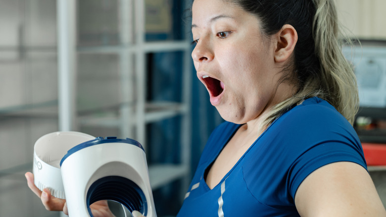 Woman with dark hair and blue shirt opening up unidentified small appliance with a look of surprise