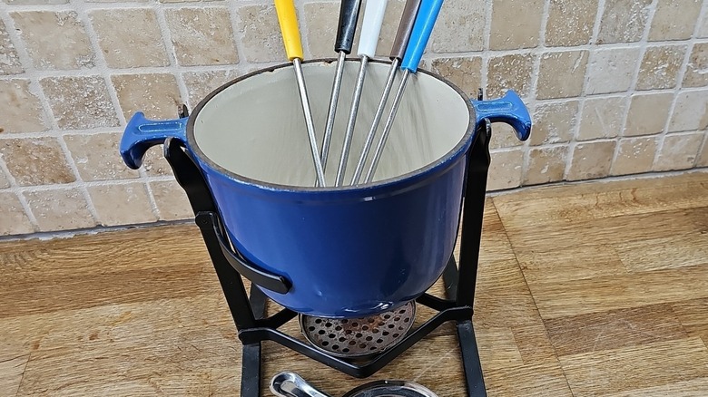 A vintage blue enamel cast iron Le Creuset fondue set sits on a wooden kitchen countertop with a ceramic tile backsplash.