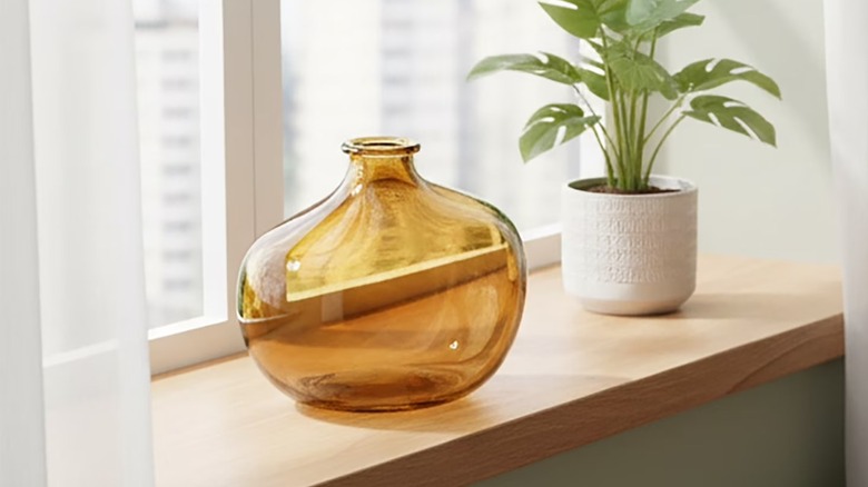 An round amber glass vase sits on a window sill near a potted plant.