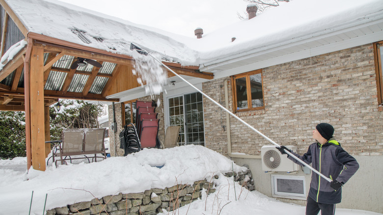 A teenage boy pushes snow off a roof with a rake