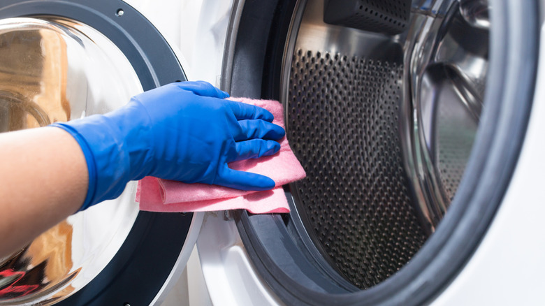 A hand wearing a blue cleaning glove and holding a pink rag wiping down the seals of a washer