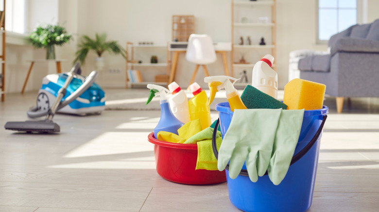 A red and a blue bucket full of cleaning supplies and a vacuum in a living room with furniture in the background