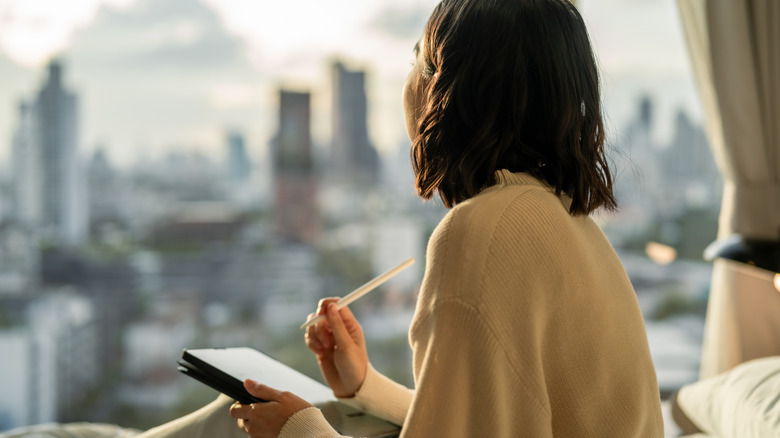 A woman sitting on her bed, looking out the window, with a notebook and a pen in her hand