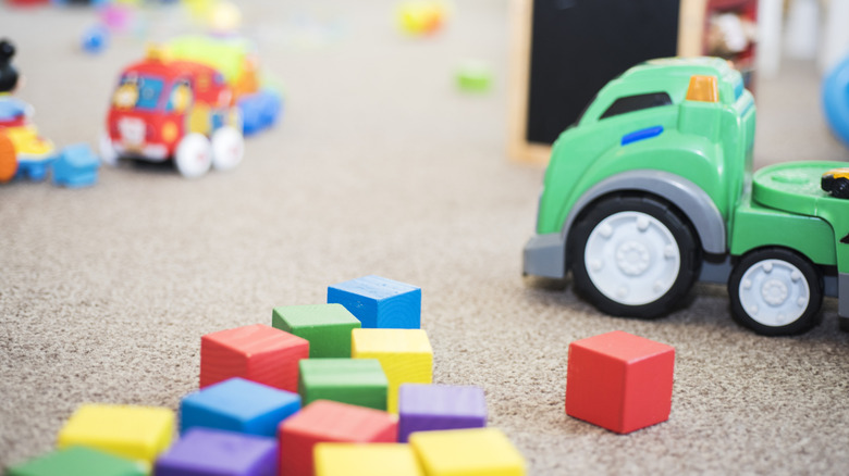 A close-up of a messy play area, with wooden blocks, plastic trucks, and other toys lying around on a carpeted floor
