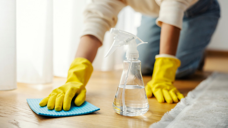 A person on their hands and knees with jeans, a cream sweater, and yellow cleaning gloves scrubbing the floor with a blue sponge and water bottle