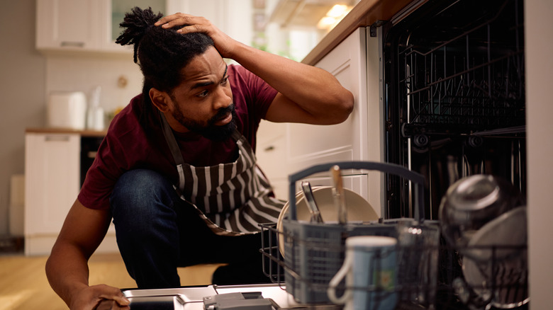 A man crouched before an open dishwasher, looking frustrated