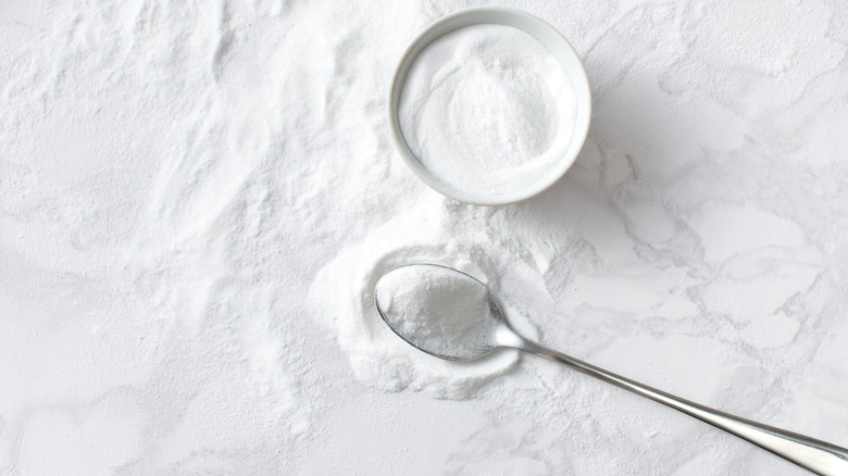 A white countertop sprinkled with baking soda that's holding a spoon and ramekin of baking soda