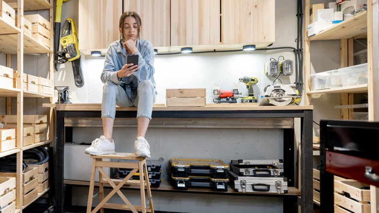 Woman looking at her phone while sitting on the workbench in a small garage.