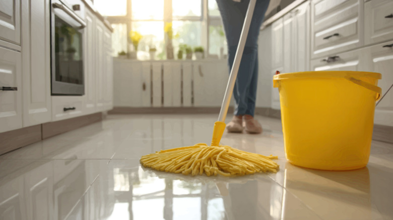 A woman is cleaning a modern kitchen floor with a mop and bucket. The floor is highly reflective, showing sunlight streaming in.