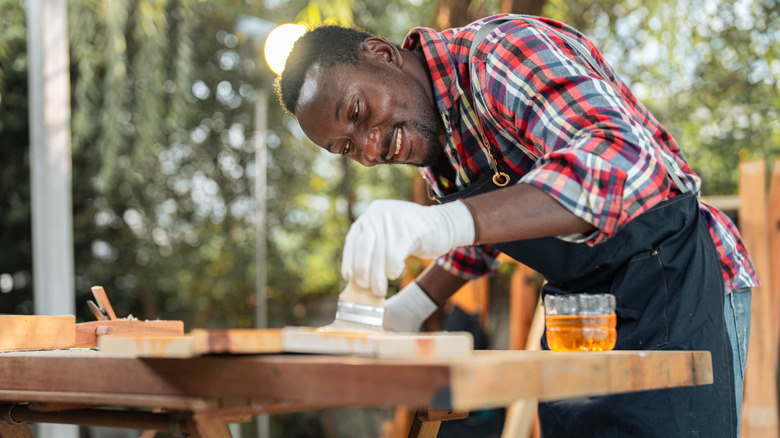 A man applies stain to a piece of wood with a brush