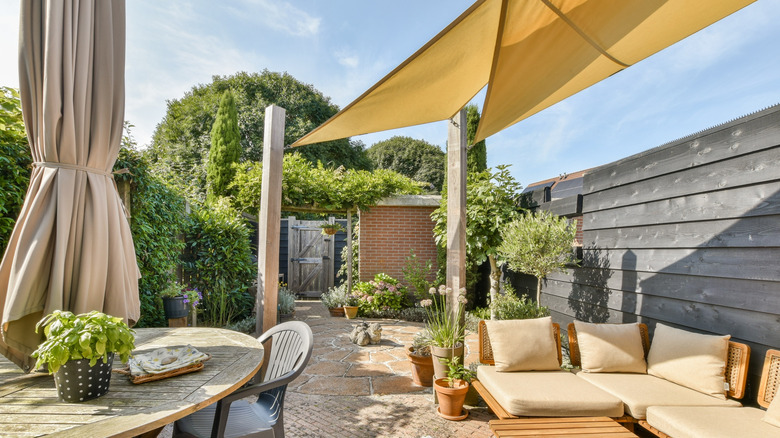 A backyard patio with chaise furniture and a round table with chairs and umbrella and a background of lush greenery.