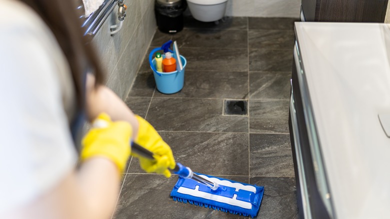 A person wearing yellow gloves mopping their tile floor in their bathroom with a bucket of cleaning products in the background