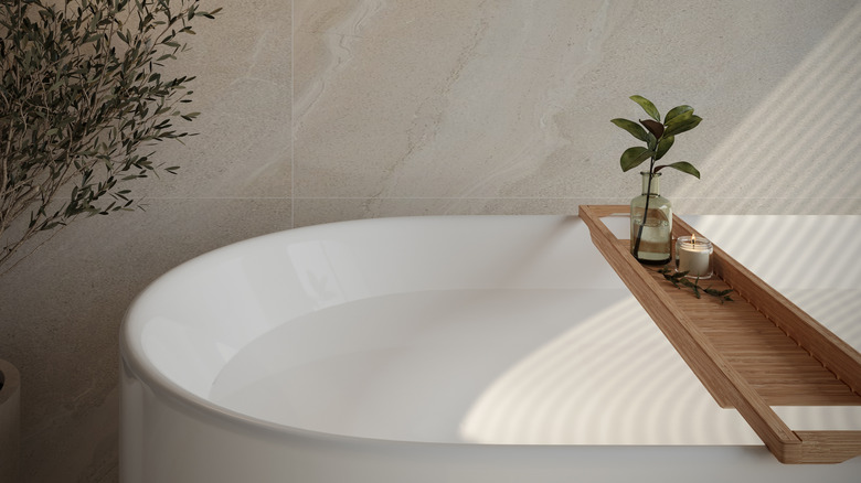 Close up of a bathtub with a bamboo tray holding a candle and a glass bottle with a sprig of green foliage
