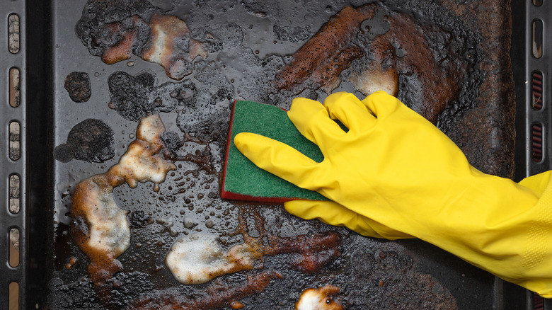 A gloved hand scrubs at a dirty baking sheet.