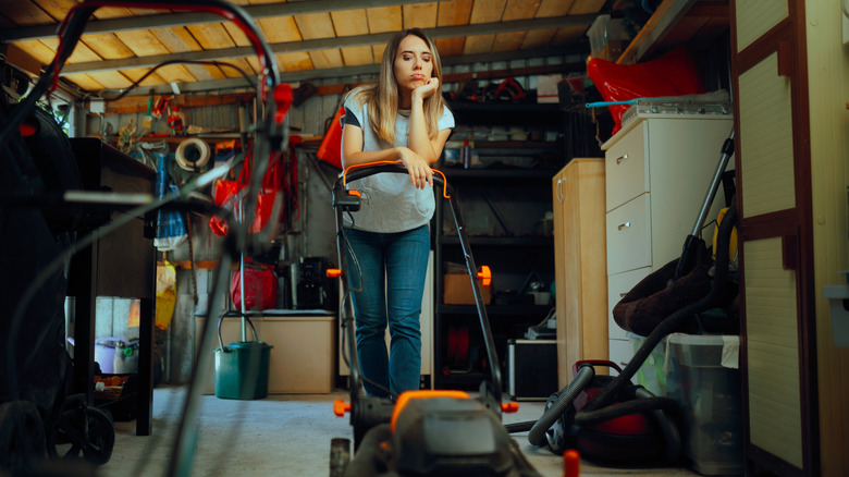 A woman looking down at a lawn mower inside a cluttered garage.