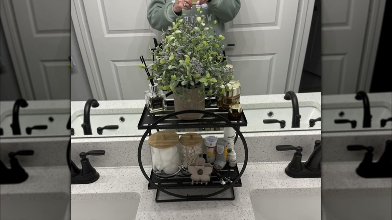the black YIWANFW shelf between two bathroom sinks, holding a potted plant and several toiletries