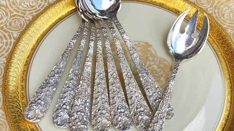 Set of Stieff sterling silver spoons in the rose pattern next to a serving fork on a gold-rimmed plate