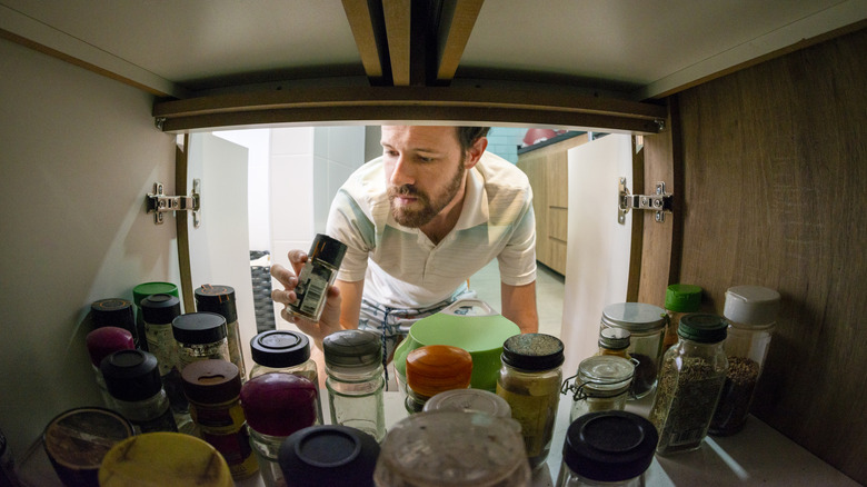 A bearded man grabs a small jar from a kitchen cabinet fill of spice jars.