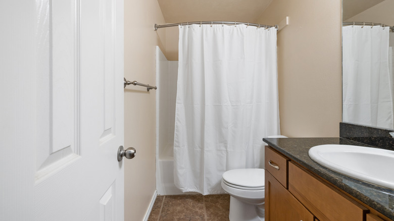 A narrow bathroom with beige walls and a white shower curtain covering a shower and bath combo.