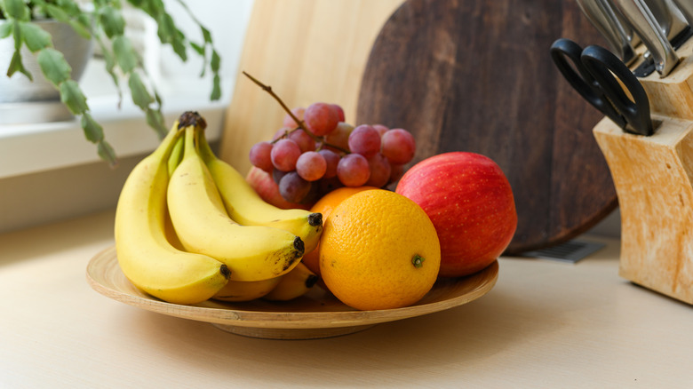 A bowl of fresh fruit on a counter, including bananas, oranges, apples, and grapes