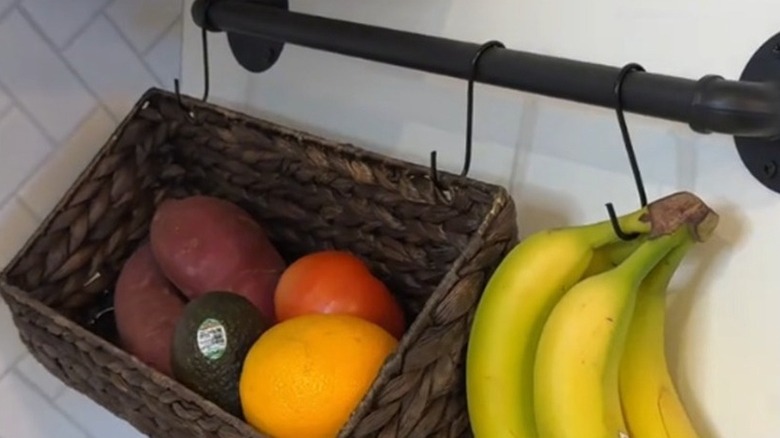 A hanging rail positioned on the wall supporting a basket with fruit, and another hook with a bunch of bananas on it