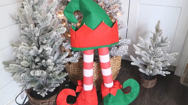 Elf legs made out of pool noodles with elf shoes and a hat next to flocked Christmas trees