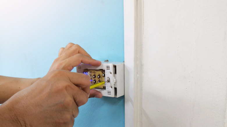 Someone using a small screwdriver to remove the faceplate of a thermostat on a blue wall.