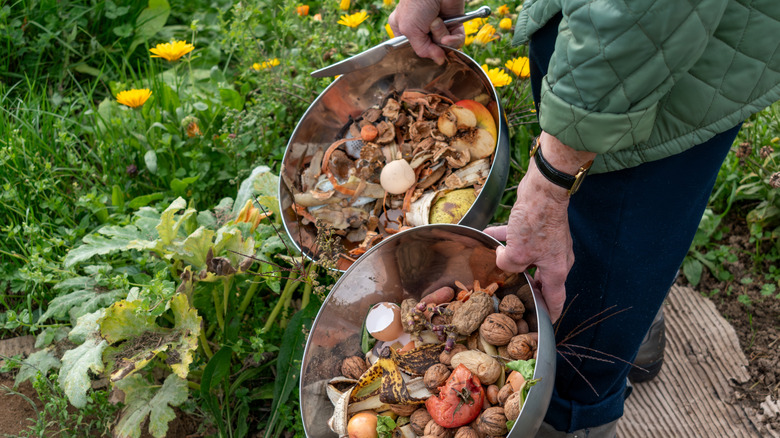 A person carries two large steel bowls full of compostable material and a small knife into their garden.