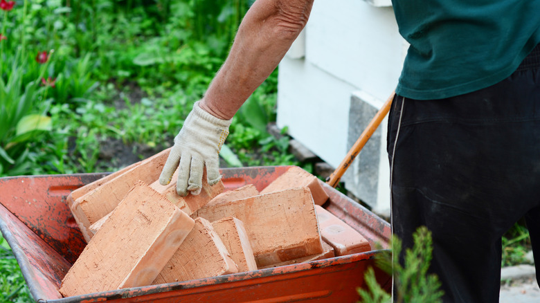 A person wearing gloves reaching into a wheelbarrow filled with bricks.