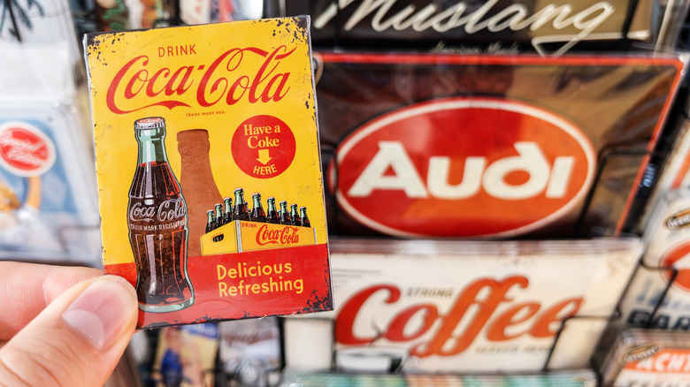 Fingers holding up a Coca Cola collectible with different brand collectibles in the background.