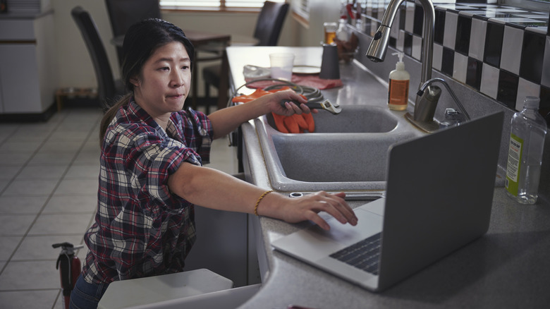 A person references their laptop while working on a kitchen sink DIY