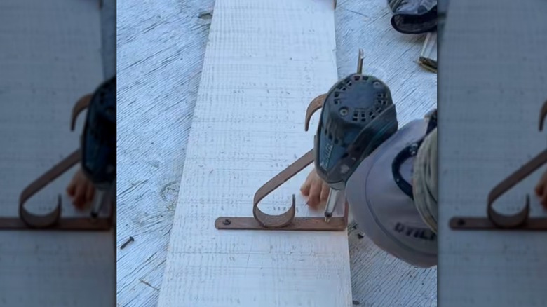 Someone using an electric screwdriver to attach a bracket to a white shelf.