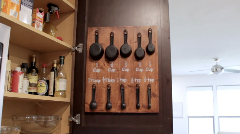 A DIY measuring cup organizer with a wooden base mounted inside a cabinet door.
