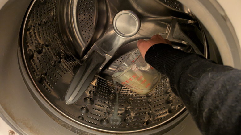 A light skinned hand pours lemon juice from a measuring cup into a front-loading washing machine.