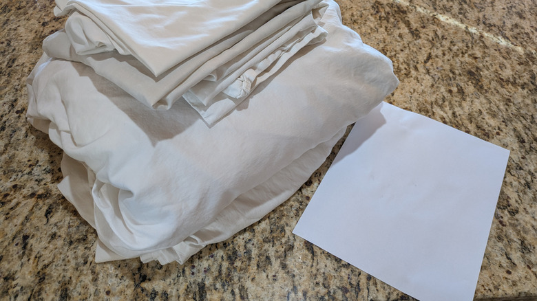 Folded white bed sheets sit next to a piece of white paper for color comparison.