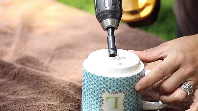 A person holds a drill bit at an angle when drilling a hole in the base of a teacup.