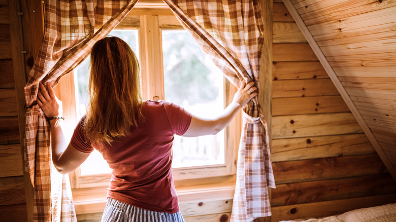 A person pushes tied-back curtains to one side as they look out the window in a wood-walled room.