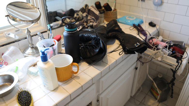Messy tile counter in bathroom covered with various items including shoe, cup, hair tools, and other miscellanea
